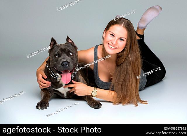 Beautiful sporty young woman lying on floor and hugging adult grey amstafford terrier dog. Studio shot over gray background. Copy space