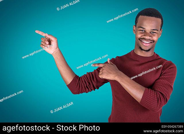 Confident African American male in red sweater cheerfully smiling and looking at camera while pointing aside with both hands against blue background