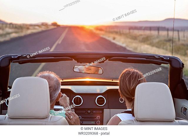 View from behind of senior couple in a convertible sports car driving on a highway at sunset in eastern Washington State, USA
