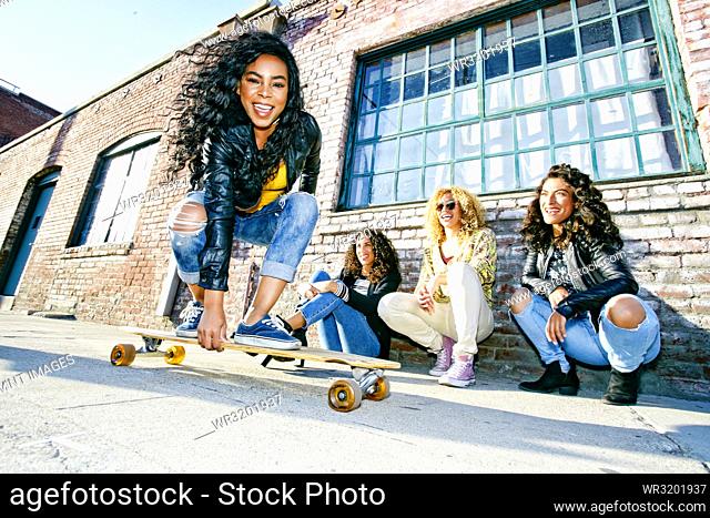 Three young women with curly hair squatting in front of old building, watching smiling young woman riding a skateboard