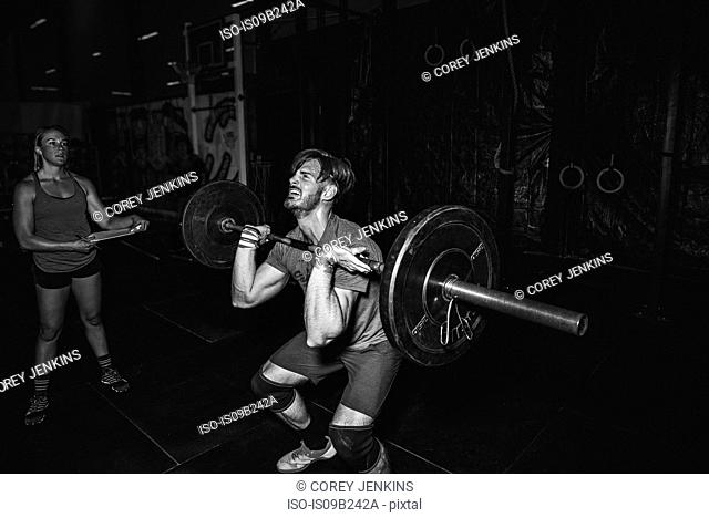 Trainer observing cross training athlete lifting barbell in gym