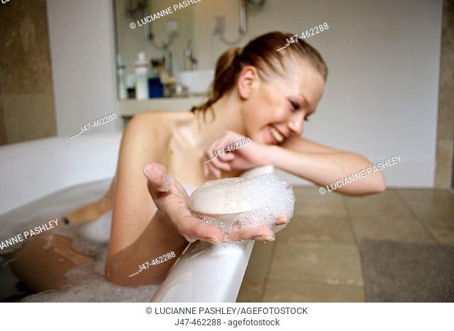 21 year old girl in the bath, leaning on the side, smiling holding a bar of soap out to the camera