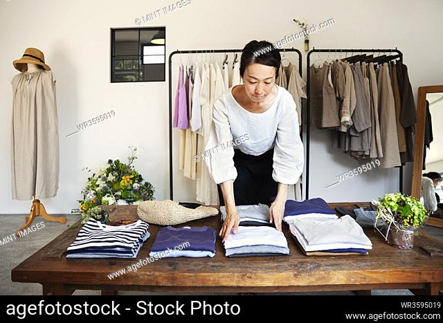 Japanese woman standing in a small fashion boutique, arranging T-Shirts on a coffee table