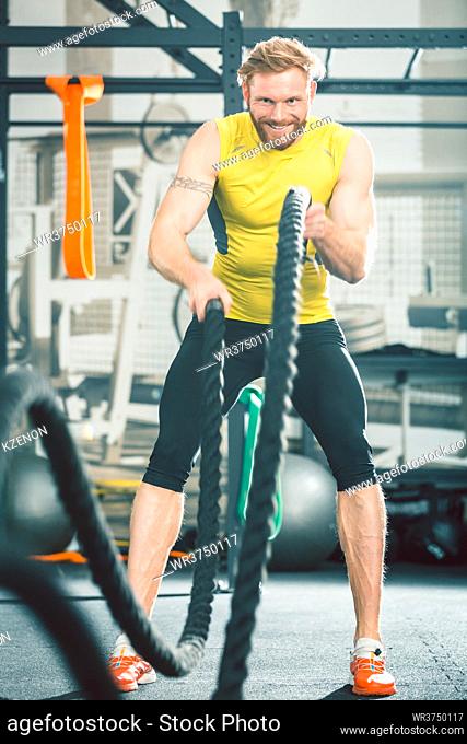Full length low angle view of a handsome bodybuilder exercising with heavy battle ropes during intense functional training workout at the gym
