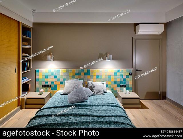 Bedroom in a modern style with a large bed with many pillows and a coverlet on the colorful tiles background. There are two wooden nightstands, glowing lamps