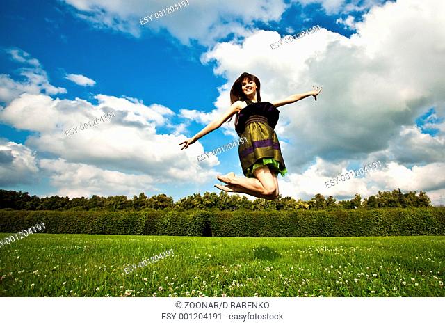 Pretty smiling girl with long brown hairs is jumping on green meadow