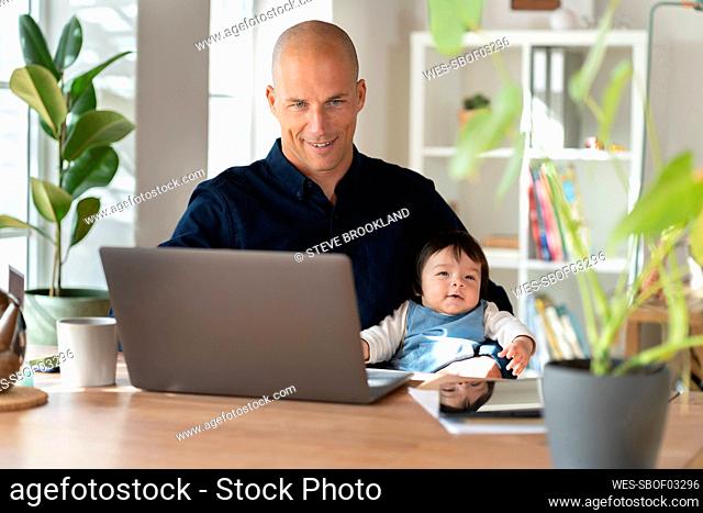 Father working on laptop while sitting with his baby in home office