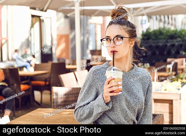 Young woman in gray sweater at street cafe drinks cocktail and looks away