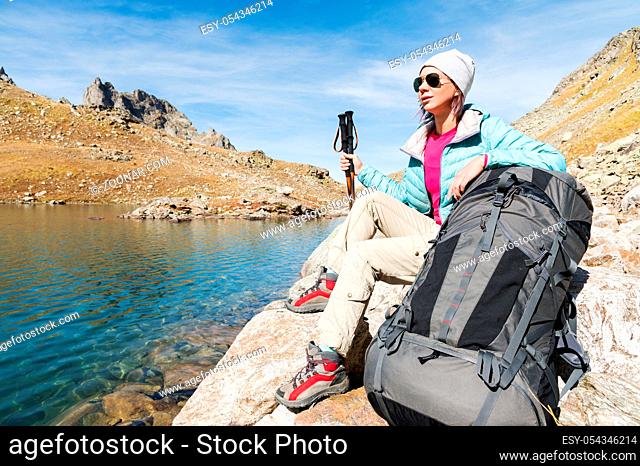 A tourist girl wearing sunglasses down jacket and hat with a backpack and mountain equipment with handles for tracking in her hands is sitting and relaxing...
