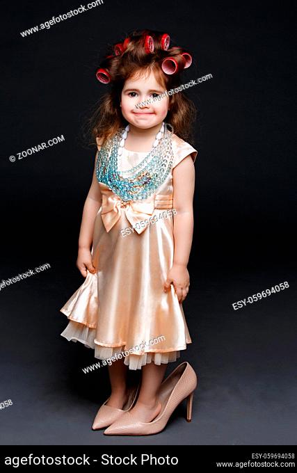 Cute little girl in dress and big high heels shoes with red lips and rollers in hair standing over dark background. Studio shot. Copy space