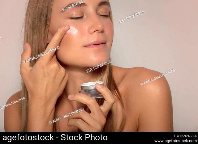 Portrait of a Beautiful Woman with Closed Eyes Applying Facial Moisturizer Cream. Natural Cosmetics. Isolated on Beige Background. Beauty Treatment