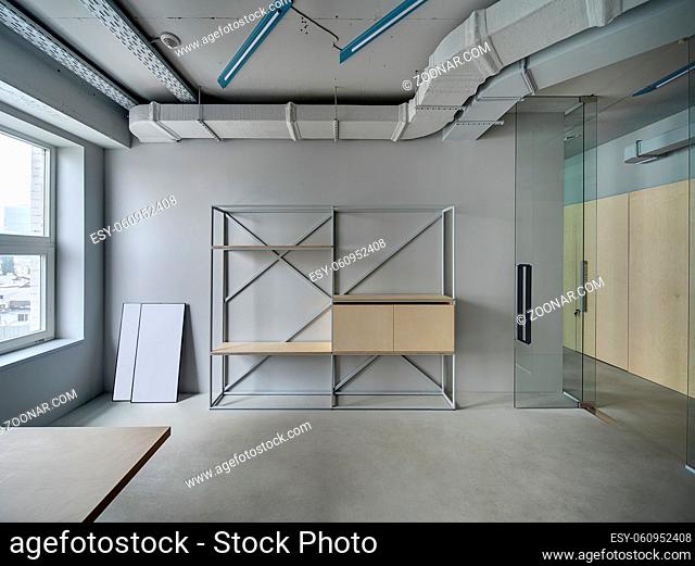 Business room in a loft style with gray walls. There is a window, a metal stand with the wooden shelves and lockers, a glass partition with a glass open door
