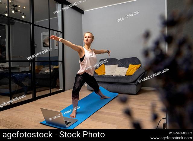 Woman with arms outstretched doing yoga on exercise mat by laptop at home