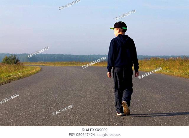 A boy in a cap and a sweatshirt goes forward in the middle of the highway in the field