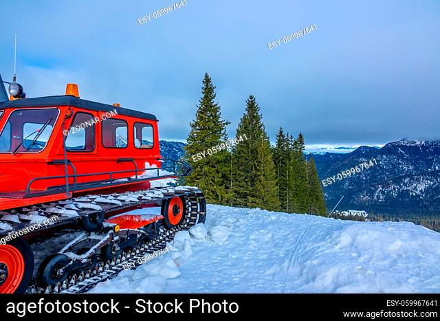 Cloudy sky over snow-capped mountain peaks. Wooded slopes. Red snowcat in the foreground