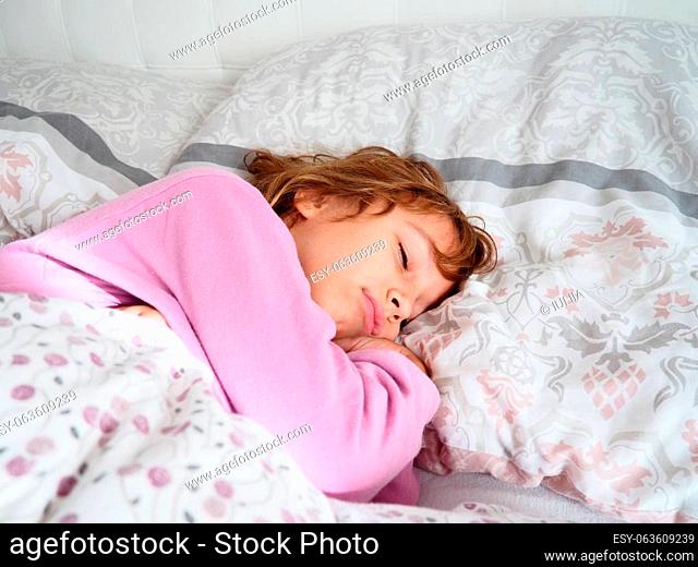 A beautiful caucasian girl of 8 years old with blond hair, dressed in pink pajamas, sleeps on a bed with a fluffy blanket, hugging a pillow