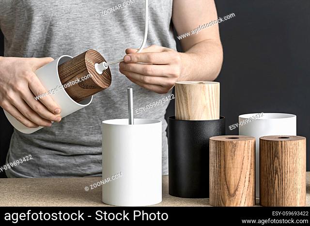 Lamp assembling in the workshop. Man holds a white lamp with a wooden part per the cable. On the table there are other lamp billets. Indoors. Closeup