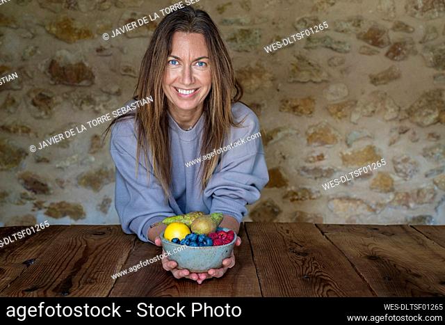 Smiling female nutritionist with various fruits in bowl sitting at table against stone wall