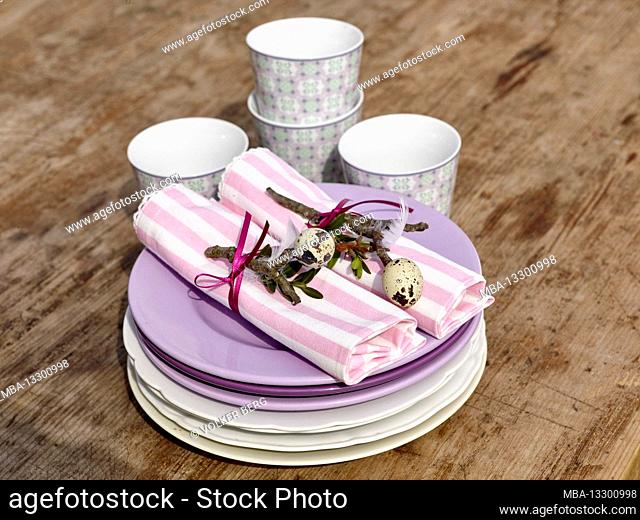 Easter breakfast, two cloth napkins decorated with box twigs and quail eggs impaled on them, surrounded by a purple ribbon on a stack of plates along with four...