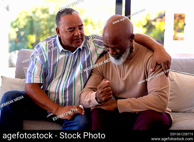 Multiracial senior man consoling male friend crying while sitting on sofa in nursing home
