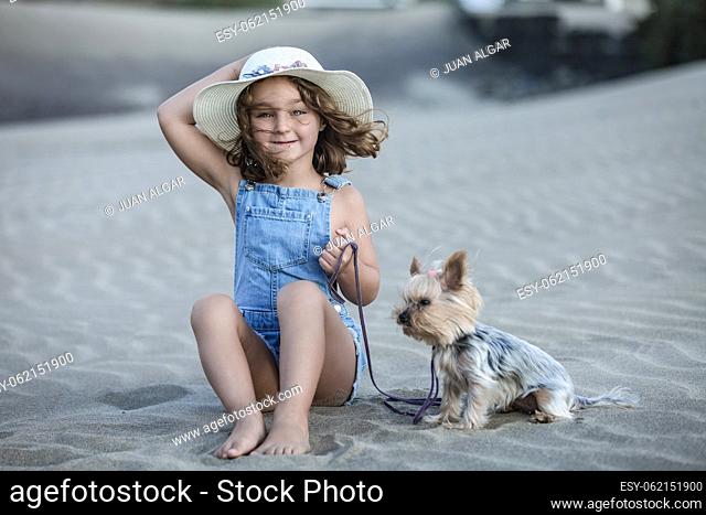 Beautiful girl with hat looking straight ahead sitting on the beach sand holding the leash of her pet yorkshire terrier