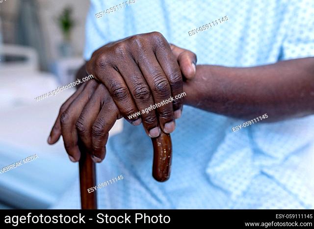 Midsection of african american male patient sitting on hospital bed holding cane