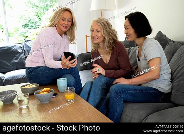 Mid-shot of middle age woman showing photos on her phone to two female friends while sitting on sofa at home