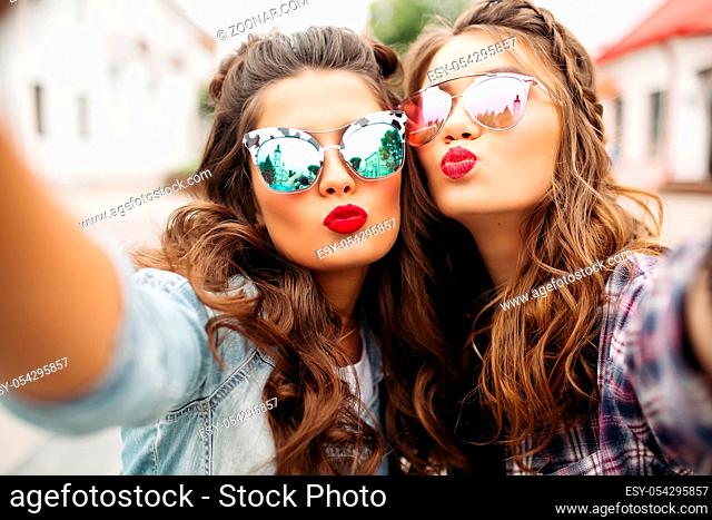Portrait of two ladies with red lips and wavy hair making selfie wearing stylish mirrored sunglasses with duck faces in the street
