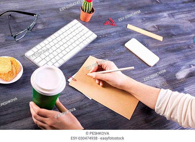 Top view of woman's hands holding coffee cup and writing on brown paper placed on wooden desktop with blank white smart phone, keyboard, cactus and other items