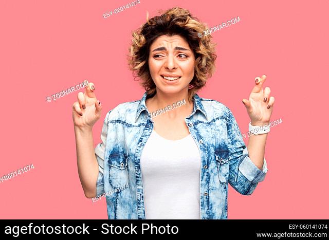 Portrait of hopeful young woman with curly hairstyle in casual blue shirt standing with crossed fingers, clenching teeth and looking away with worry face