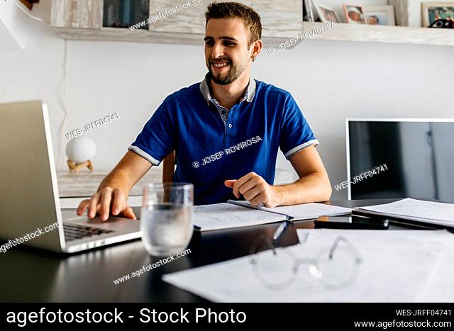 Smiling handsome young man using laptop while doing homework at table