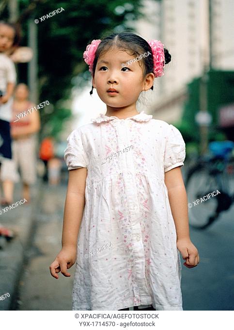 A young 3 to 5 yr old Chinese girl with her hair tied in two pink buns standing on a street in Shanghai, China