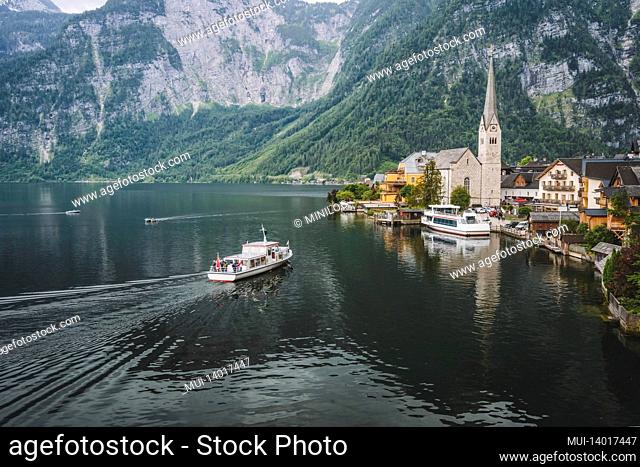 mountain lake and tourist boat reaching hallstatt village with alps in background. austria