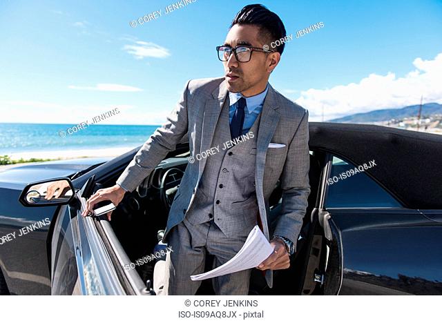 Young businessman getting out of car at coastal parking lot with paperwork and smartphone