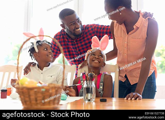 Happy african american parents with son and daughter wearing bunny ears painting colourful eggs