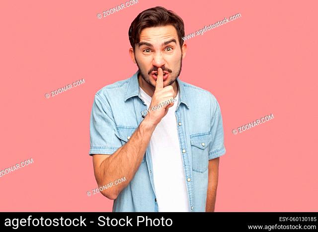Portrait of serious or angry bearded young man in blue casual style shirt standing with silence sign gesture, finger on lips and looking at camera