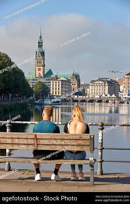 20 August 2023, Hamburg: A young couple sits on a park bench at the Binnenalster. In the background you can see the city hall and the Reesendamm bridge at the...