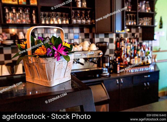 A closeup view of colorful flowers arranged in a small wooden basket inside a coffeeshop. Beautiful decor with industrial coffee machine blurred in background