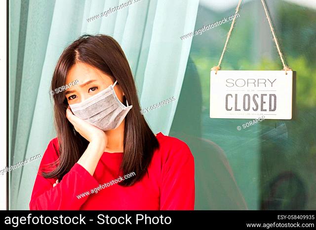 Asian young woman wear face mask protect she sad notice sign wood board label ""SORRY WE ARE CLOSED PLEASE COME BACK AGAIN"" hanging through glass door front...