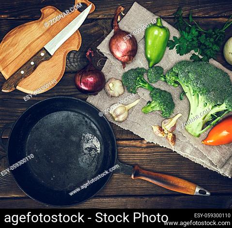 empty round black cast-iron frying pan and fresh vegetables on a brown wooden table, top view