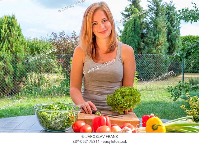 Beautiful young woman is preparing vegetable food outdoors. She is holding a head of green leaf lettuce looking at the camera