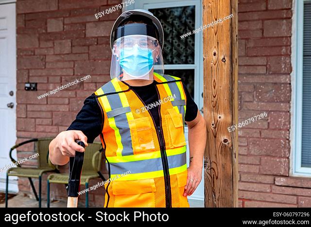 Medium close shot of a man wearing an orange fluo visibility jacket, covid protective face mask and plastic visor leaning on the handle of a shovel