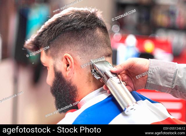 Handsome bearded man, getting haircut by barber, with electric trimmer at barbershop