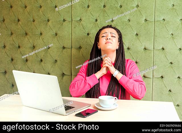 My God, please help me. Portrait of young girl freelancer with black dreadlocks in pink blouse, sitting in cafe and holding palm hands and praying