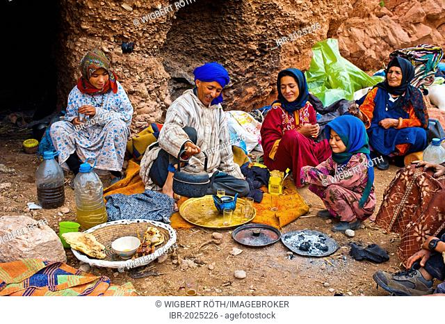 Nomadic cave-dwellers sitting in front of their cave-dwelling, a man wearing a blue turban is pouring tea into a pot on a copper tray