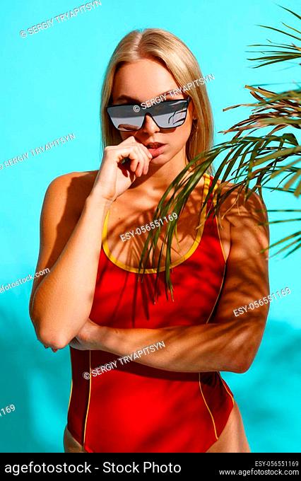 Sportive woman in red swimsuit poses on cyan background in studio. Girl in swimwear ready to take a tan. Model with slim body in swimming underwear