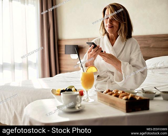 Smiling blond senior woman using smart phone while sitting with breakfast in hotel room