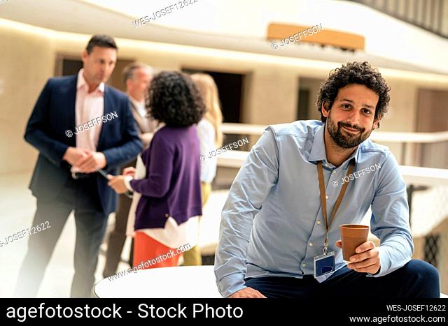 Smiling businessman holding coffee cup with colleagues discussing in background