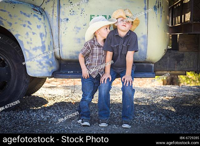 Two young boys wearing cowboy hats leaning against an antique truck in a rustic country setting