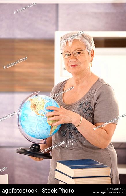 Senior teacher standing at desk, holding globe, teaching geography in elementary school